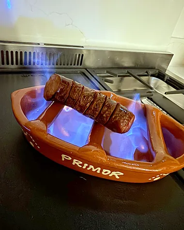 A whole scored chouriço flaming over blue fire in a terracotta assador